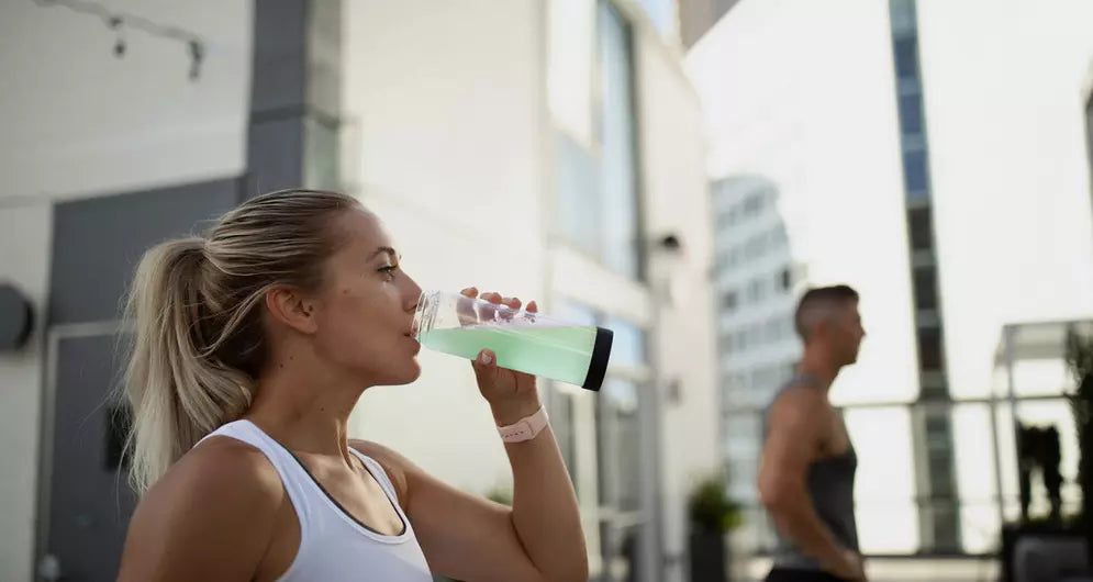 Woman in activewear drinking a clear green protein shake from a shaker bottle outdoors in an urban fitness setting.