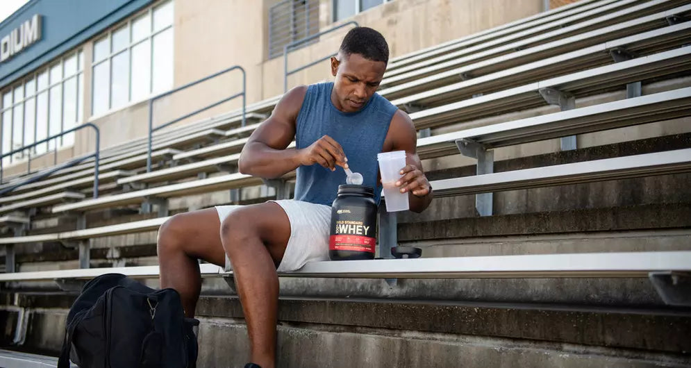 Man bereitet Whey-Proteinshake auf Stadiontribüne vor, mit Becher und Löffel in der Hand