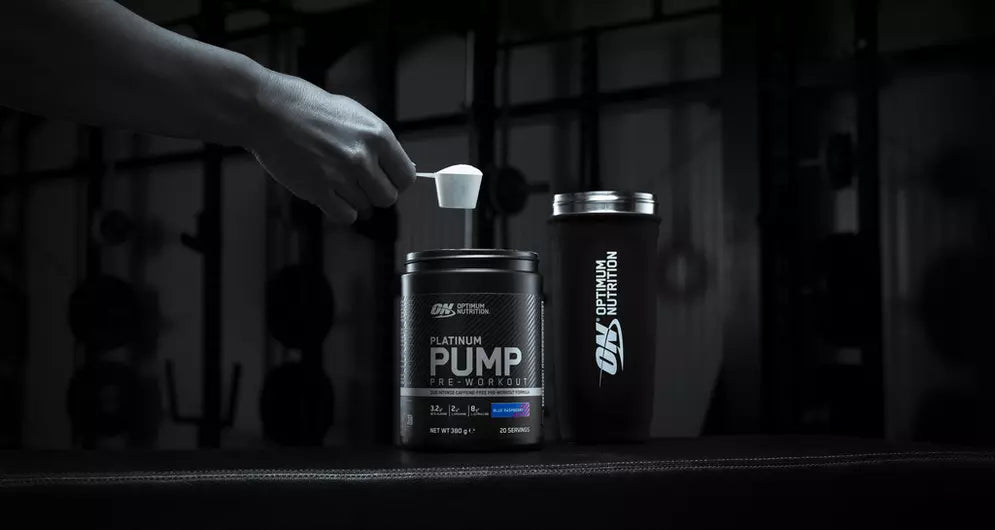 Hand scooping Platinum Pump pre-workout from a tub with Optimum Nutrition shaker nearby in a gym setting.