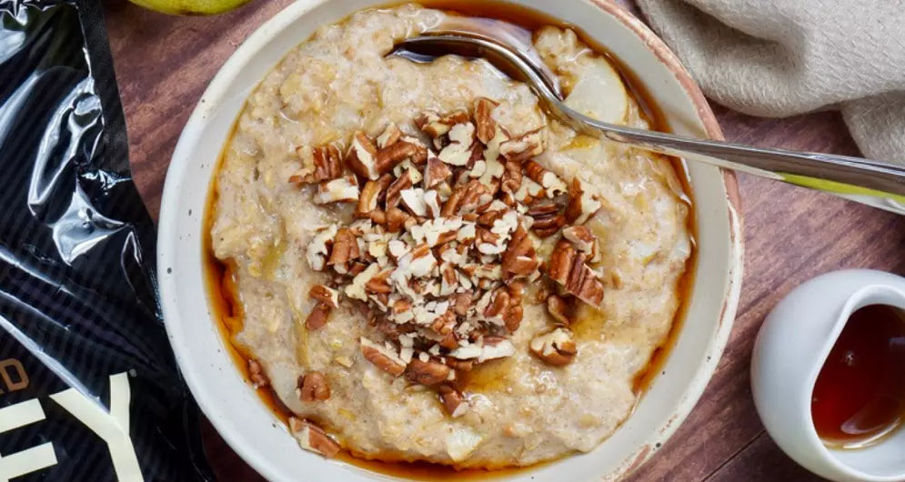 Spiced vanilla, pear, and maple porridge topped with chopped pecans, served in a bowl with a spoon, alongside protein powder and maple syrup.