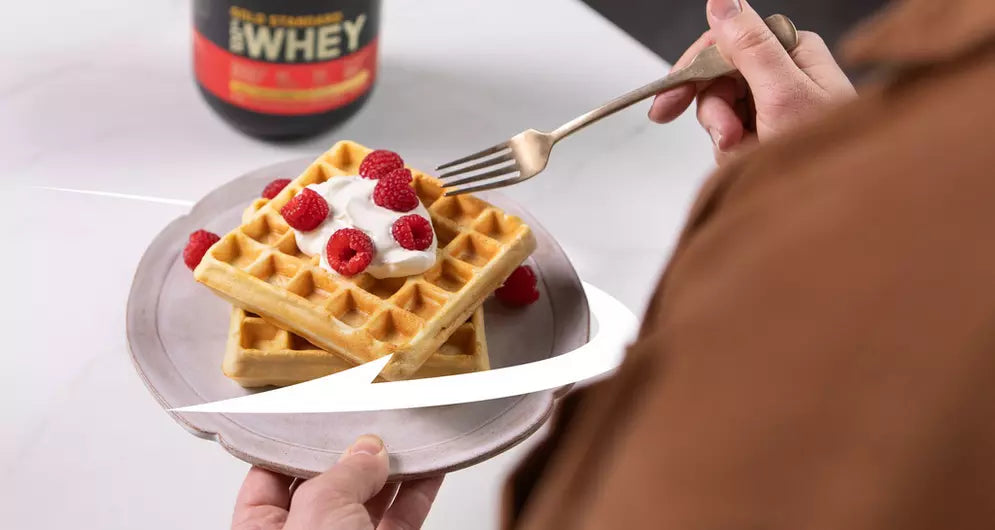 Vanilla protein waffles topped with yogurt and raspberries on a plate, with a hand holding a fork and a whey protein tub in the background.