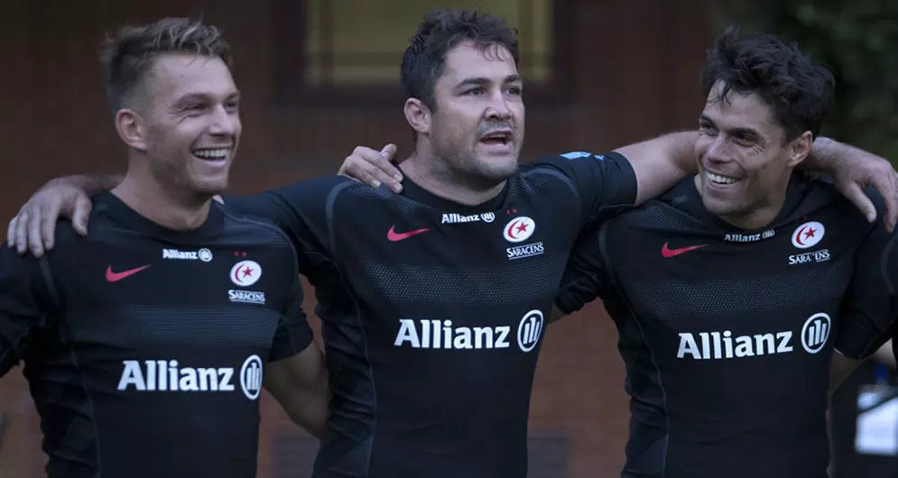 Three rugby players in black Allianz Saracens kits standing arm in arm, smiling after training.
