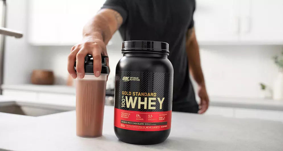 Hand holding a shaker bottle next to a tub of Gold Standard 100% Whey protein powder on a kitchen counter.