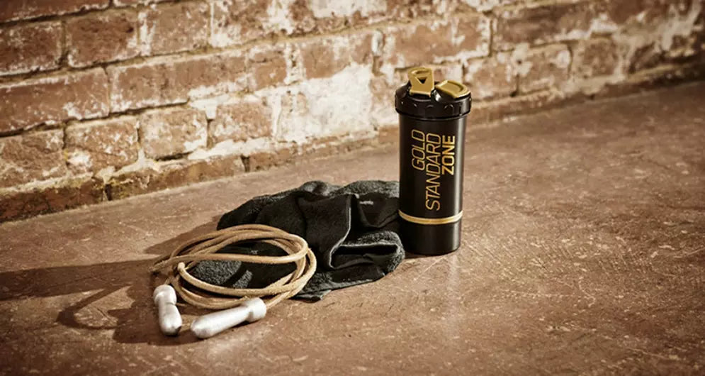 Gold Standard shaker on a gym floor with a jump rope and towel, placed against a rustic brick wall.
