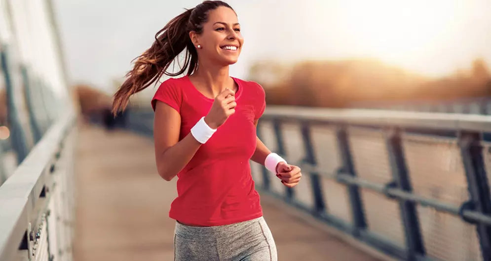 Smiling woman jogging outdoors on a bridge at sunrise, wearing a red top and grey leggings.