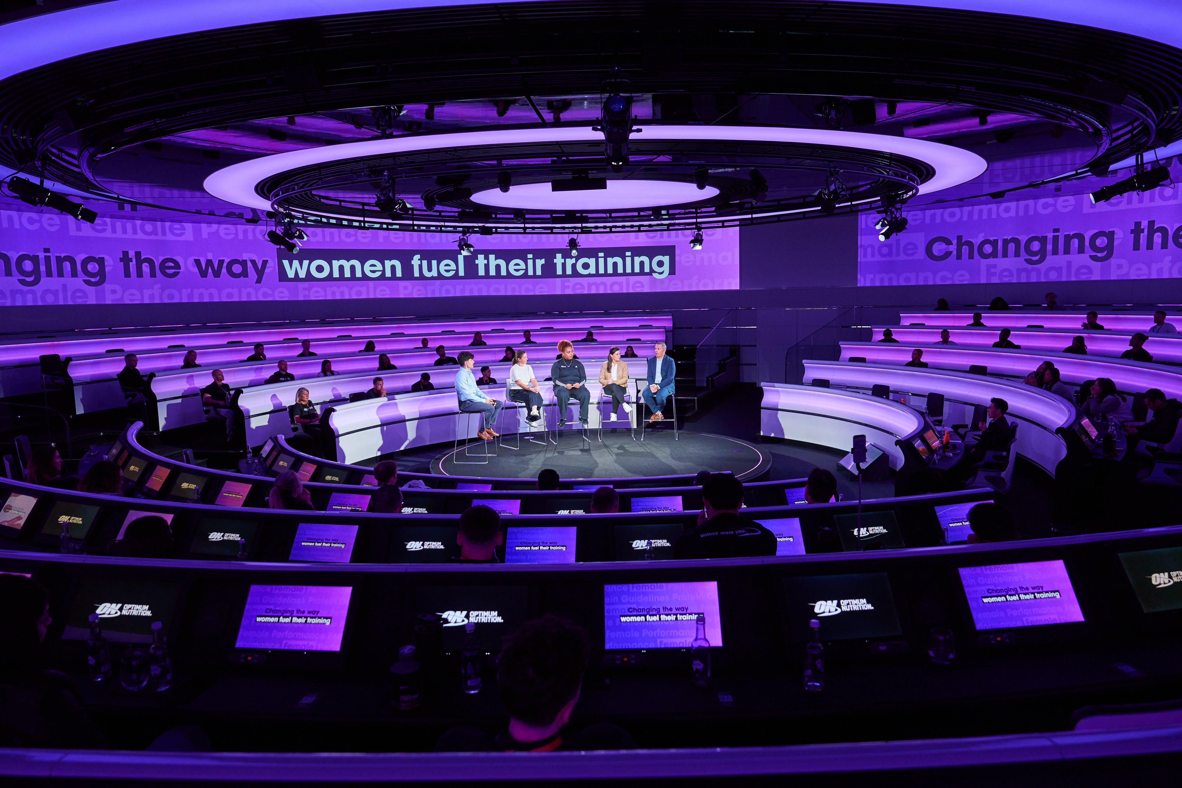 Panel discussion in a circular auditorium with multiple screens displaying text about women's training and performance