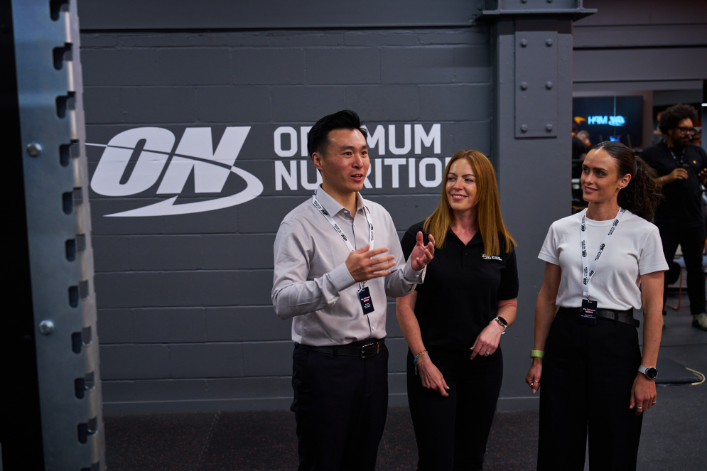 Optimum Nutrition logo displayed in a gym setting with three people standing in front of it