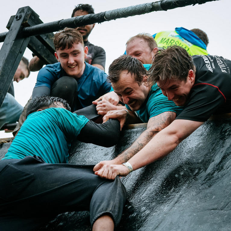 Group of people helping each other climb a wet obstacle during an outdoor challenge event