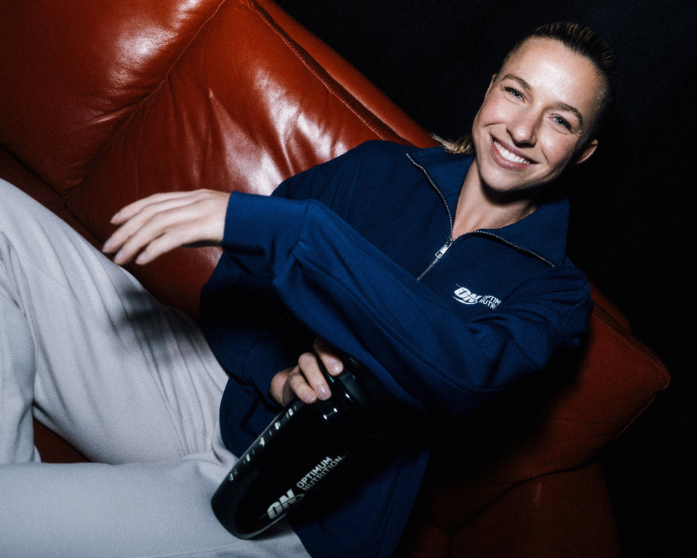 Jacky Groenen on a red leather couch holding a black shaker with a visible Optimum Nutrition logo