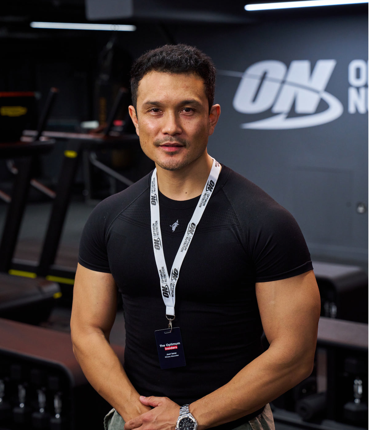 A person wearing a black shirt and a lanyard with an ON badge stands in a gym with Optimum Nutrition branding in the background