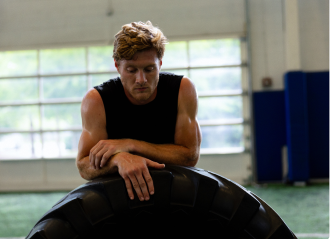 Man resting on a large tire in a gym setting, possibly after a high-intensity workout