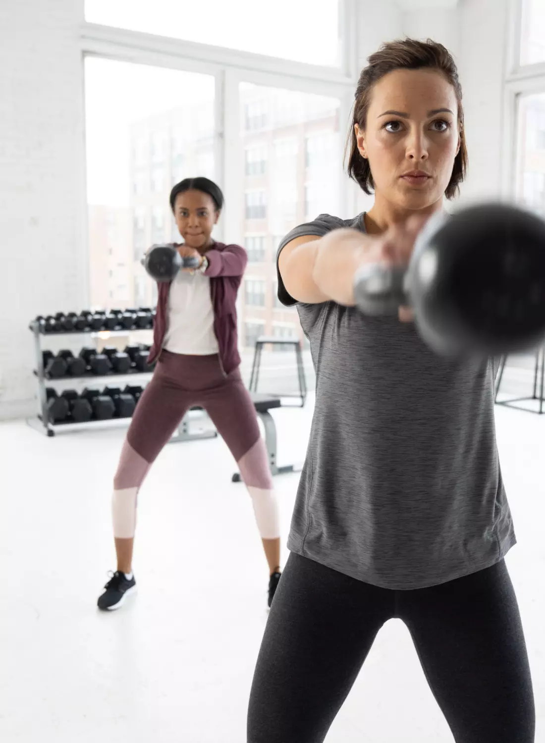 Two women performing kettlebell exercises in a gym setting with dumbbells in the background