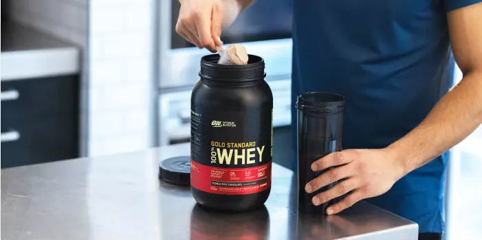 Optimum Nutrition Gold Standard 100% Whey Protein, 2.73 kg container, with a person scooping powder into a shaker