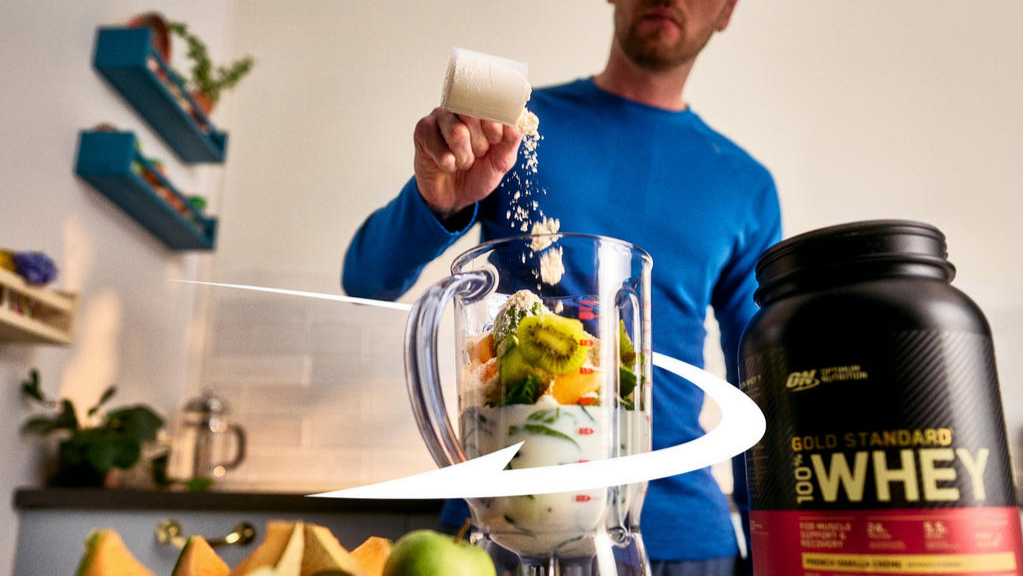 Man adding protein powder to a blender with fruits, next to a container of Gold Standard 100% Whey protein powder