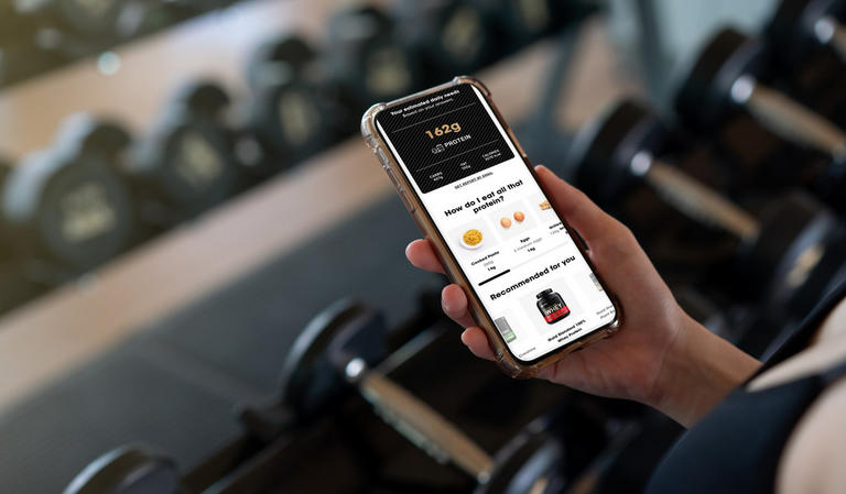 Person holding a smartphone displaying a protein goal calculator app, with gym equipment in the background