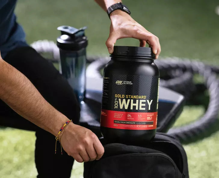 Gold Standard 100% Whey protein powder container being placed into a gym bag, next to a shaker bottle
