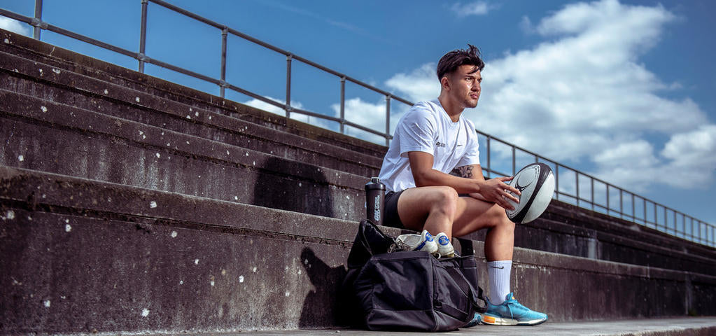 A person sitting on outdoor steps with a sports bag containing a rugby ball and athletic gear