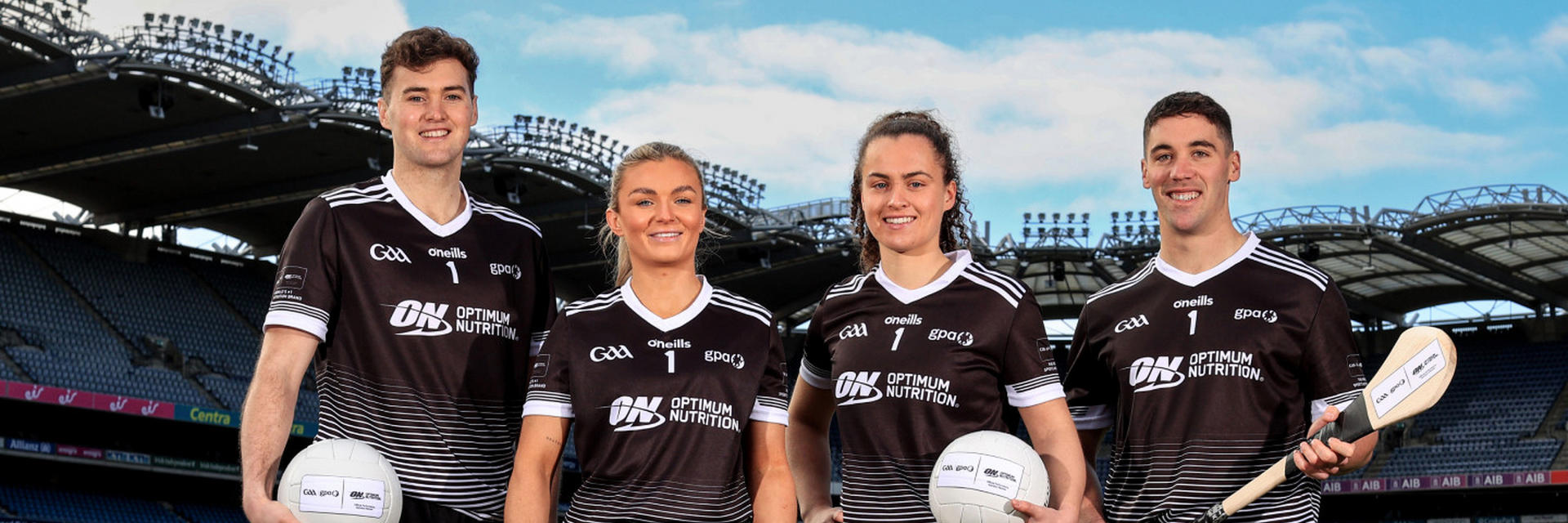 Four athletes in sports uniforms holding Gaelic footballs and a hurling stick, featuring Optimum Nutrition branding