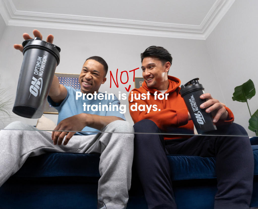 Two people sitting on a couch holding Optimum Nutrition shaker bottles, emphasizing protein use beyond training days