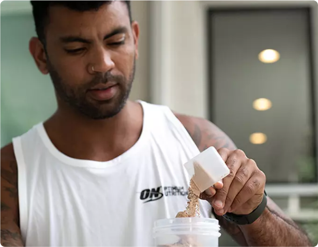 Man in gym attire pouring protein powder into a shaker bottle, wearing a tank top with "ON" logo