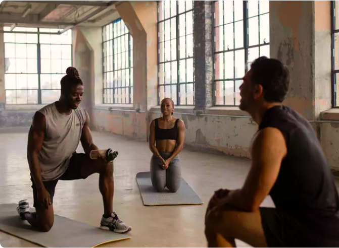 People exercising in a gym, with two individuals kneeling on mats and one standing, holding a water bottle