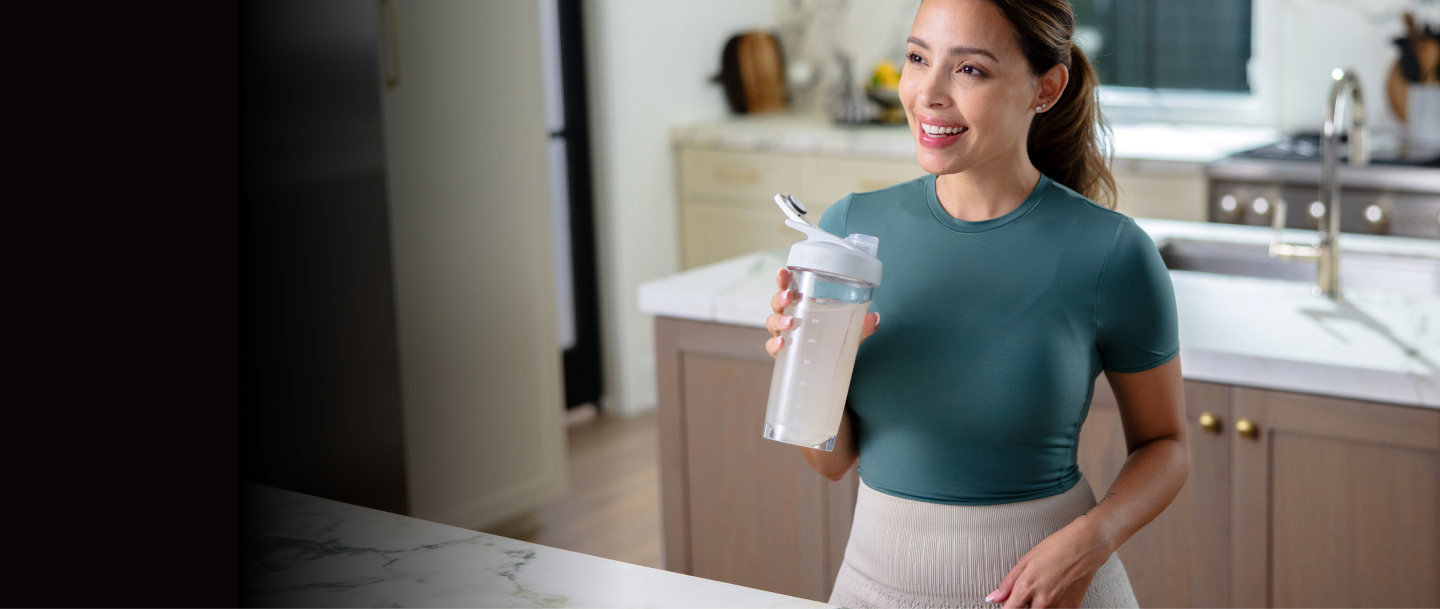 Woman holding a shaker bottle, possibly containing an amino energy drink or protein supplement, in a kitchen setting