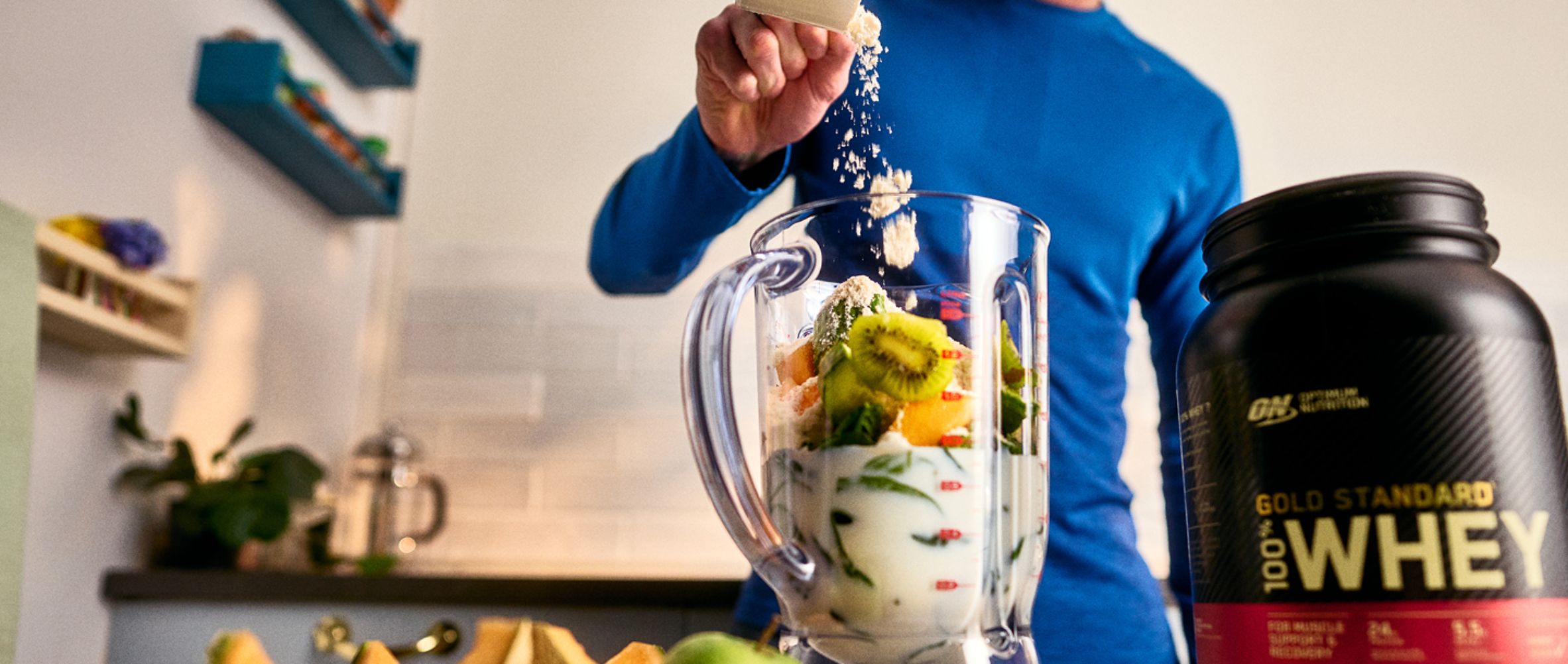 Person adding powder to a blender with fruit, next to a container of Gold Standard 100% Whey protein powder