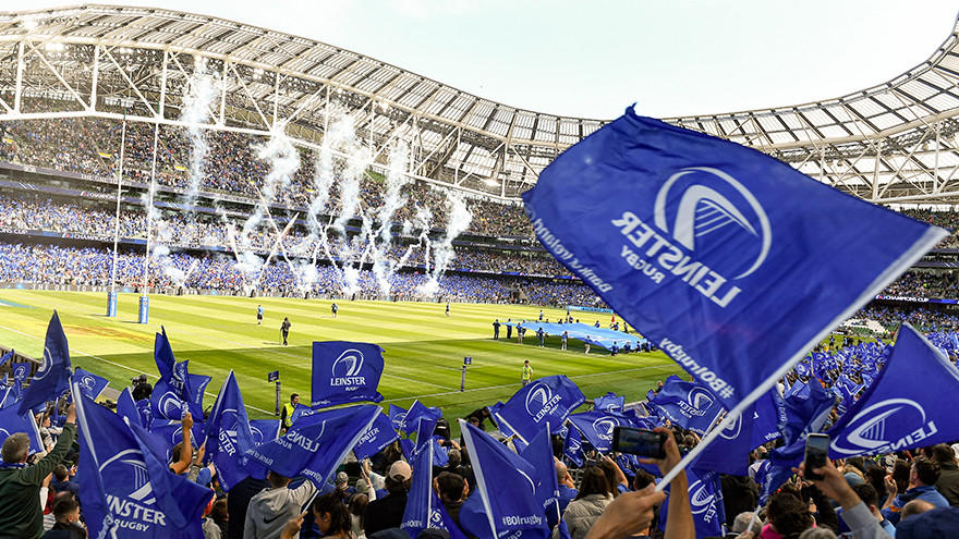 A crowd waving Leinster Rugby flags at a stadium during a match, with fireworks in the background