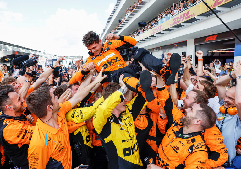 A group of people celebrating and lifting a person in orange racing attire at a motorsport event