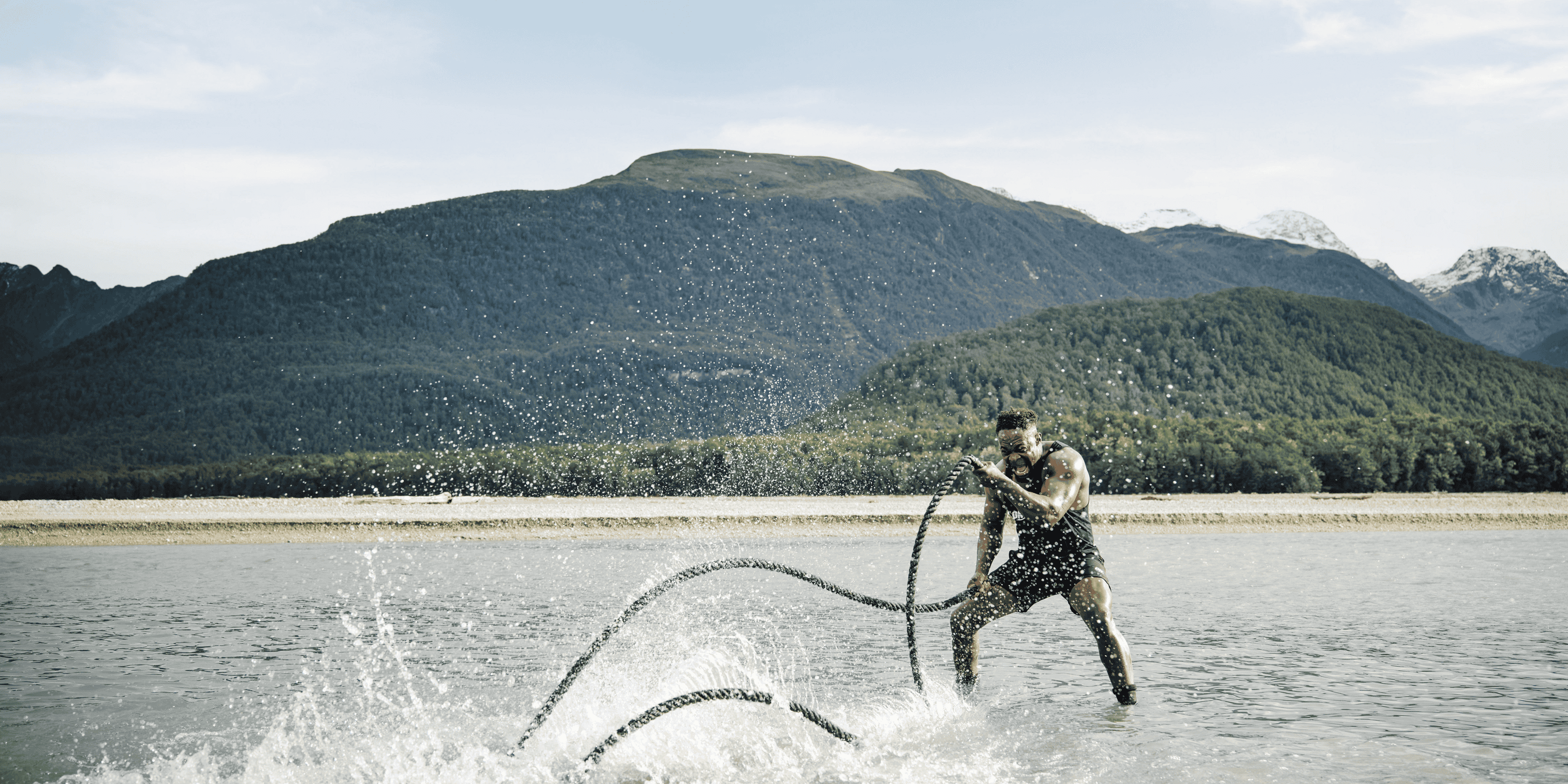 A person exercises with battle ropes in a shallow body of water, with mountains in the background