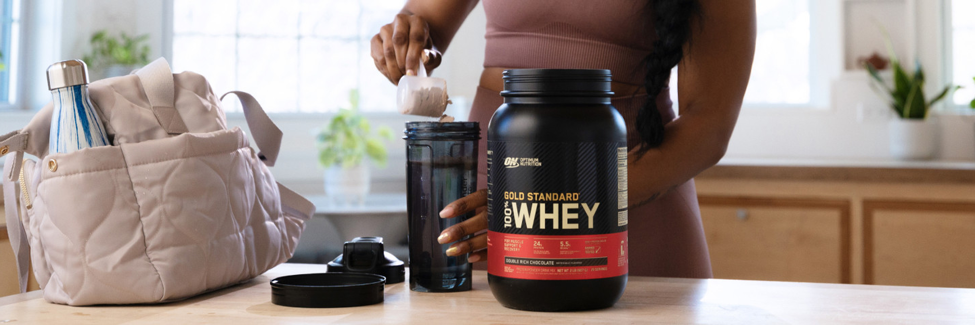 Gold Standard 100% Whey protein powder container on a kitchen counter with a person scooping powder into a shaker bottle