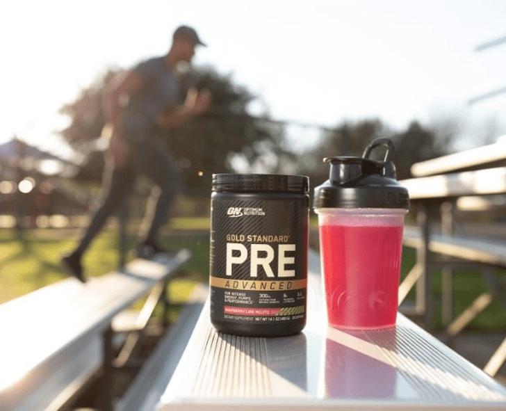 Gold Standard Pre-Workout supplement container next to a shaker bottle with a pink drink, placed on outdoor bleachers