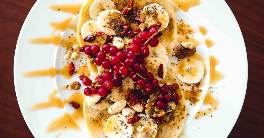 Plate of pancakes topped with sliced bananas, red currants, nuts, and a drizzle of syrup