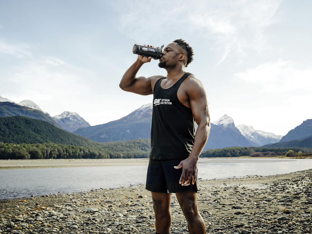 Man drinking from Optimum Nutrition shaker bottle, wearing Optimum Nutrition tank top, standing by a river with mountains in the background