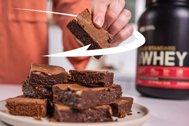 Plate of chocolate brownies with a hand reaching for one, next to a container of Gold Standard Whey protein powder