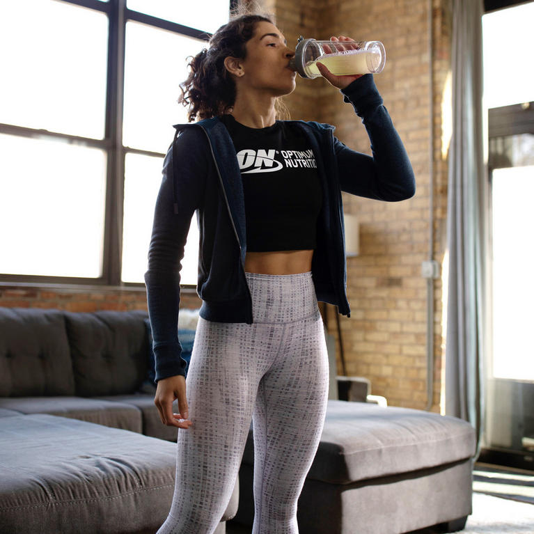Person drinking a protein shake, wearing an Optimum Nutrition shirt, in a living room setting