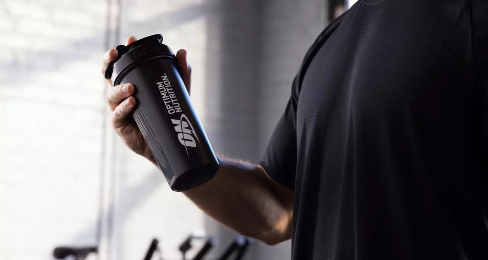 Close-up of a person holding a black Optimum Nutrition shaker bottle in a gym setting.