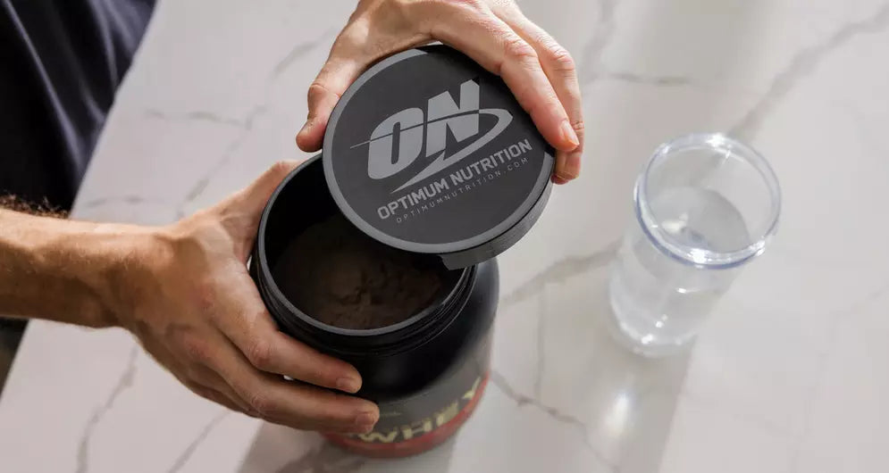 Hands opening a tub of Optimum Nutrition protein powder next to a glass of water on a kitchen counter.