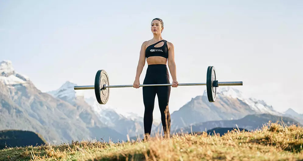 Female athlete lifting a barbell outdoors with mountains in the background, showcasing strength and power.