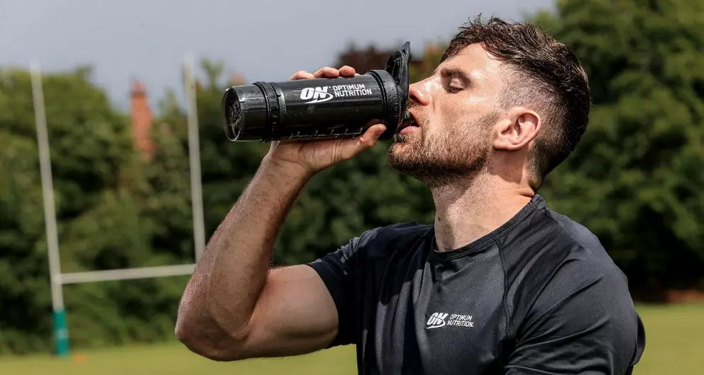 Male athlete drinking from an Optimum Nutrition shaker outdoors on a rugby field, post-training hydration or recovery.