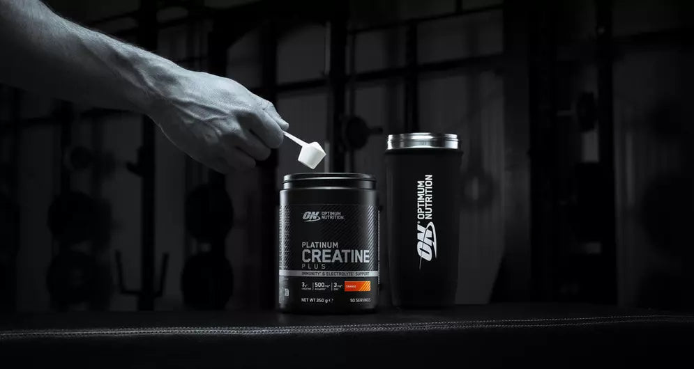 Hand scooping Optimum Nutrition creatine powder in a gym setting with ON shaker placed beside the tub.