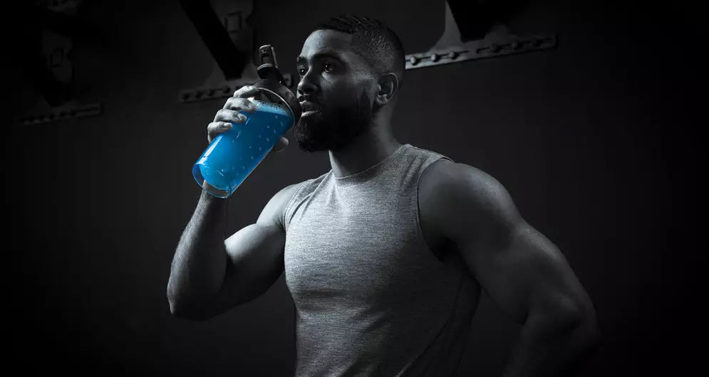 Man in grey tank top drinking a bright blue pre-workout drink from a shaker bottle in a dark gym.