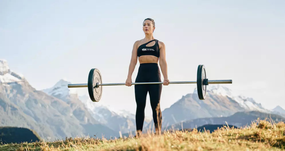 Frau hebt Langhantel vor Bergkulisse, trägt ON Sportswear, zeigt Krafttraining in der Natur
