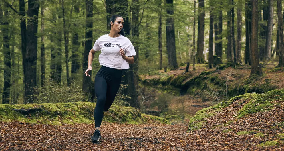 Woman running through a forest trail wearing a white Optimum Nutrition T-shirt and black leggings.