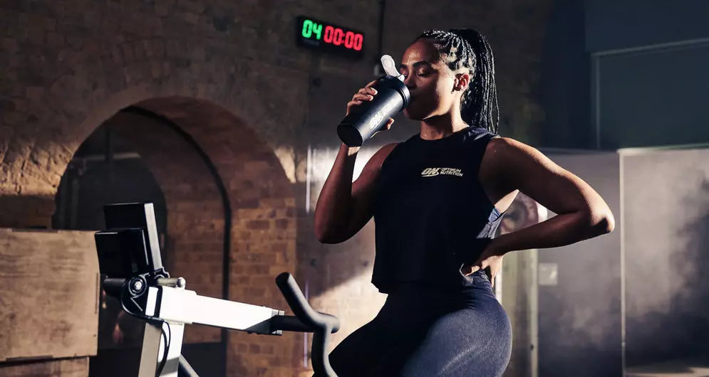Woman in gym clothes drinking from a shaker bottle while sitting on an exercise bike under a digital timer.