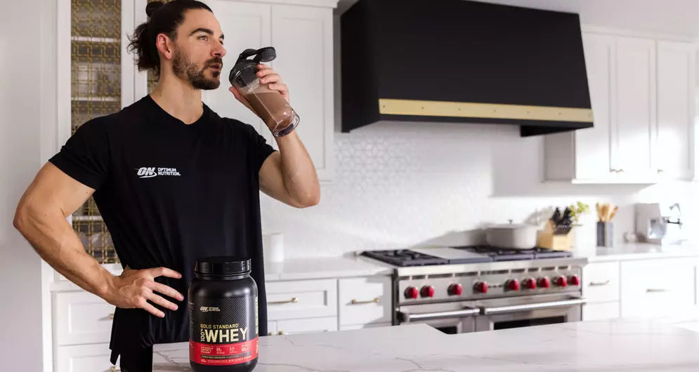 Man drinking a whey protein shake in a modern kitchen after workout, with Optimum Nutrition tub on the counter.