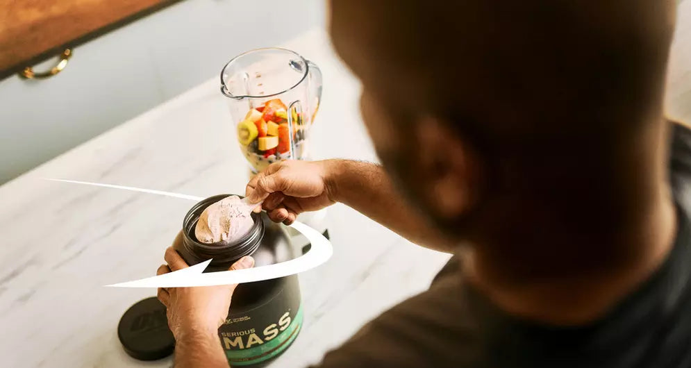 Man scooping Serious Mass protein powder next to a blender filled with fresh fruit.