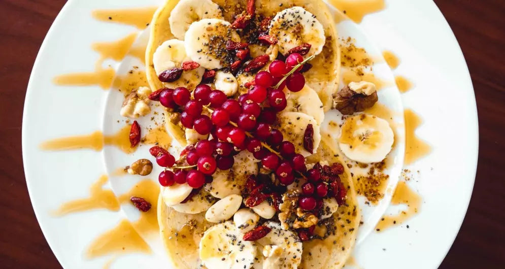 Pancakes protéinés sains garnis de banane, groseilles, noix, graines et un filet de sirop sur une assiette blanche.