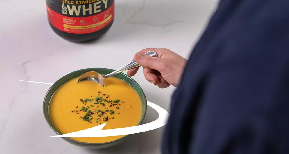 Proteinrik butternutpumpasoppa i en grön skål, garnerad med örter, med en hand som håller en sked och ett kärl med vassleprotein i bakgrunden.