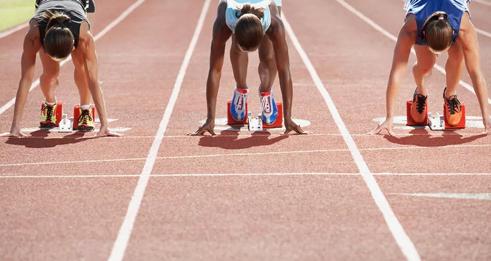 Three female sprinters in starting position on a red running track, ready for a race.